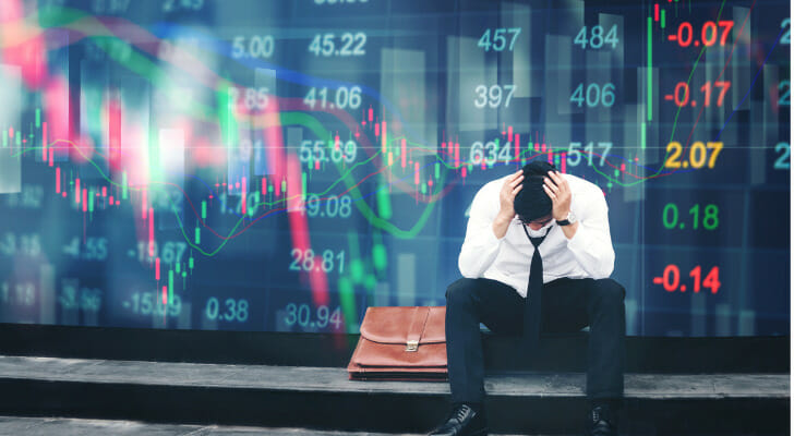 A businessman sitting with his head in his hands in front of a wall of stock prices.